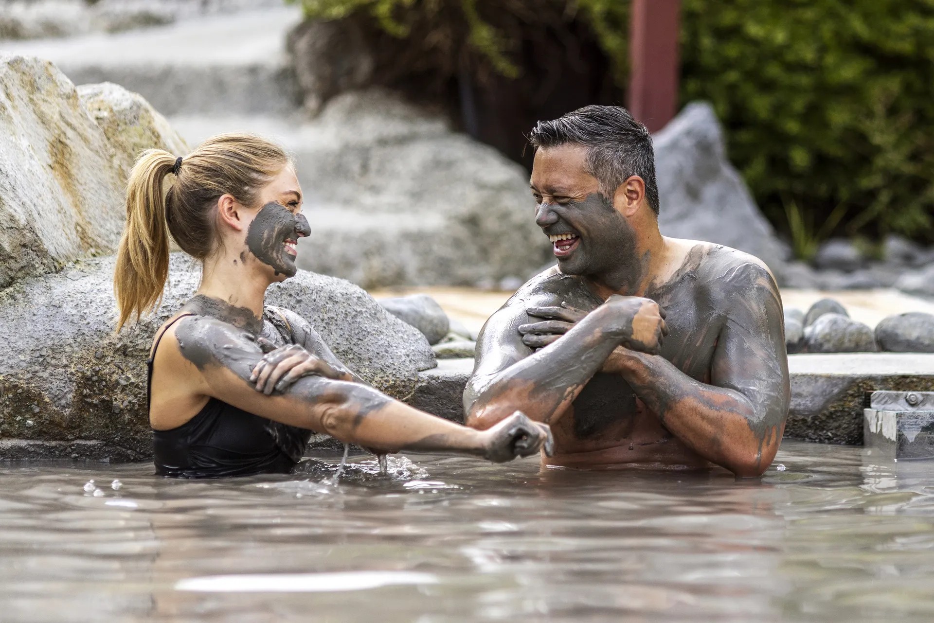 Man and woman covered in mud, enjoying an outdoor mud pool.