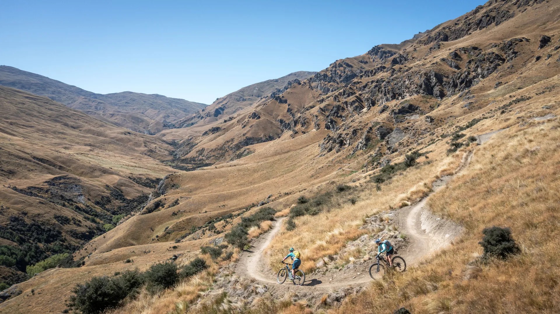 Bikers riding along a mountainous path with tussock fields and rocks.