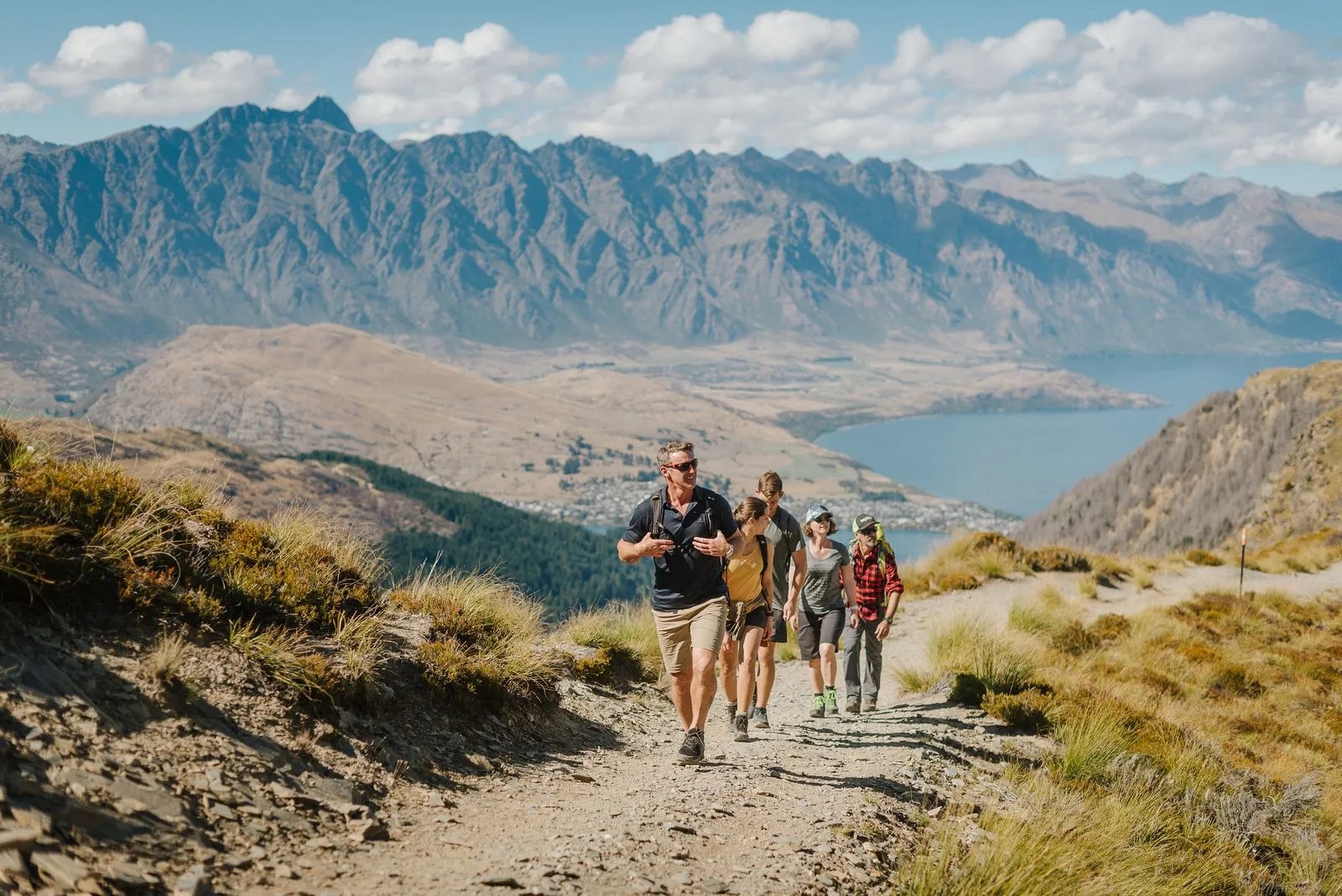 Man leading a group of walkers up a gravel mountain path, views of Queenstown and Lake Wakatipu afar.