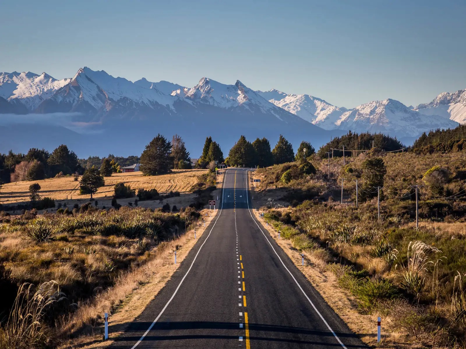 A long, straight rural road stretching through open countryside toward the snow-capped Southern Alps under a clear blue sky