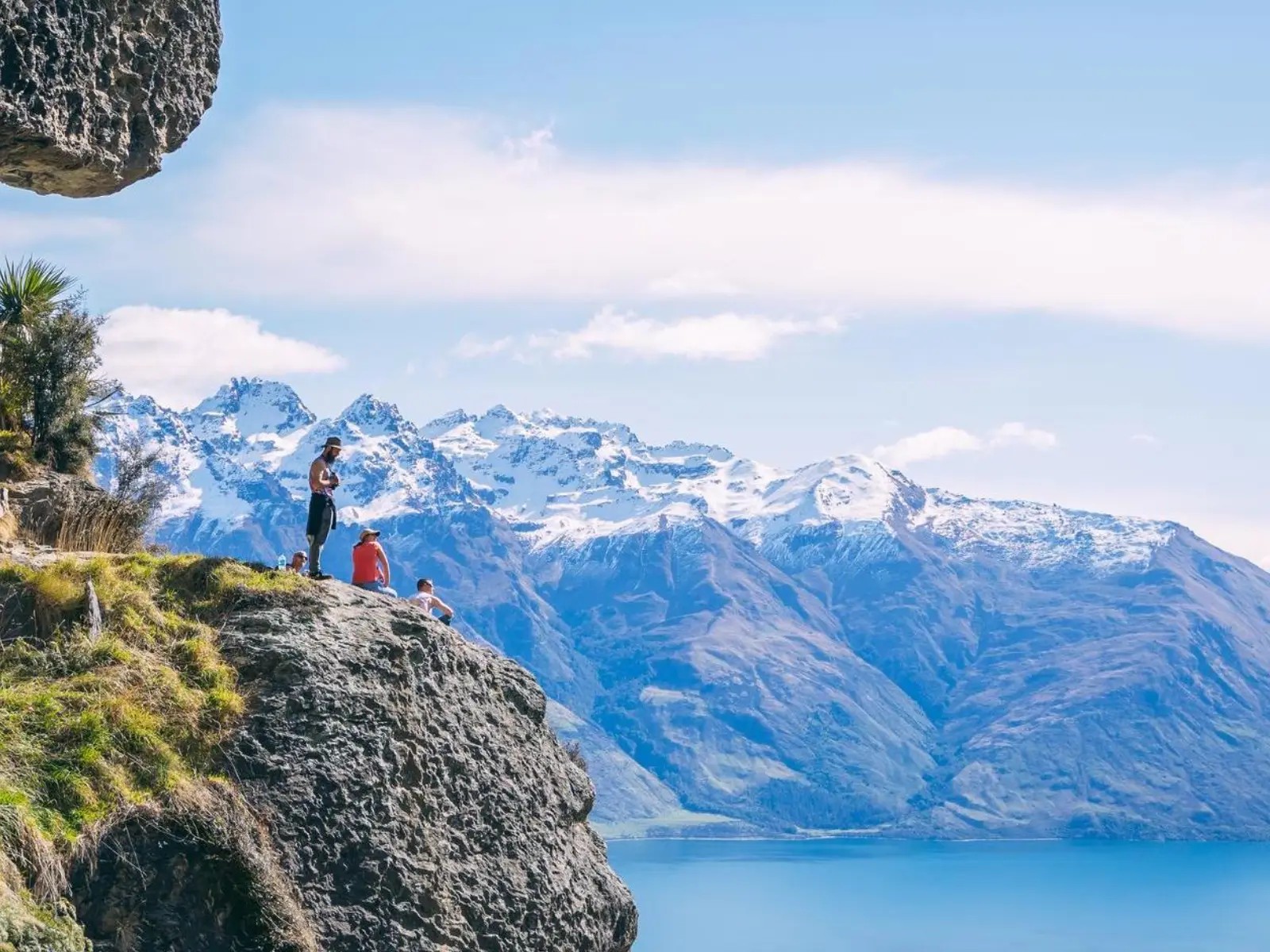 People standing on a rocky cliff overlooking a turquoise lake with snow-capped mountain ranges in the background under a clear sky
