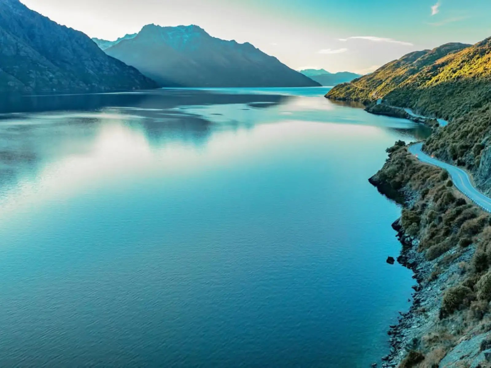 A wide view of a turquoise lake surrounded by steep mountain ranges, with a winding road tracing the shoreline under soft evening light