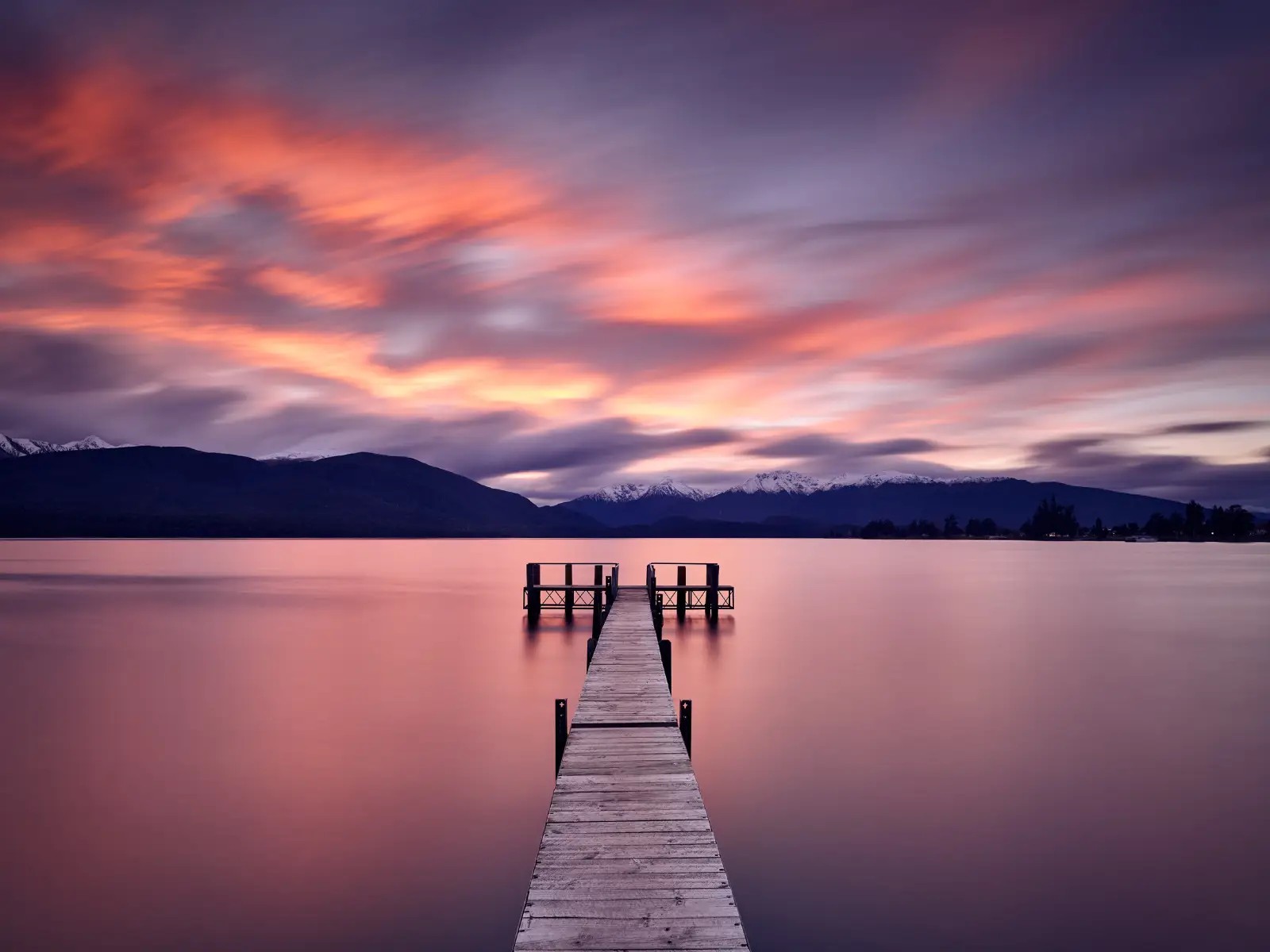 A wooden jetty extending into a calm lake at sunset, with soft pink and purple clouds reflected on the water and mountains in the distance