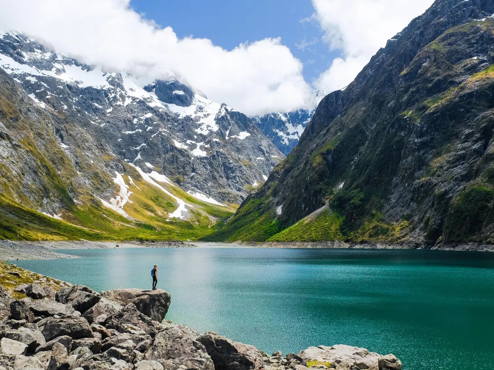 A person standing on rocky ground beside the vivid turquoise waters of Lake Marian, surrounded by steep alpine mountains with patches of snow and cloud above