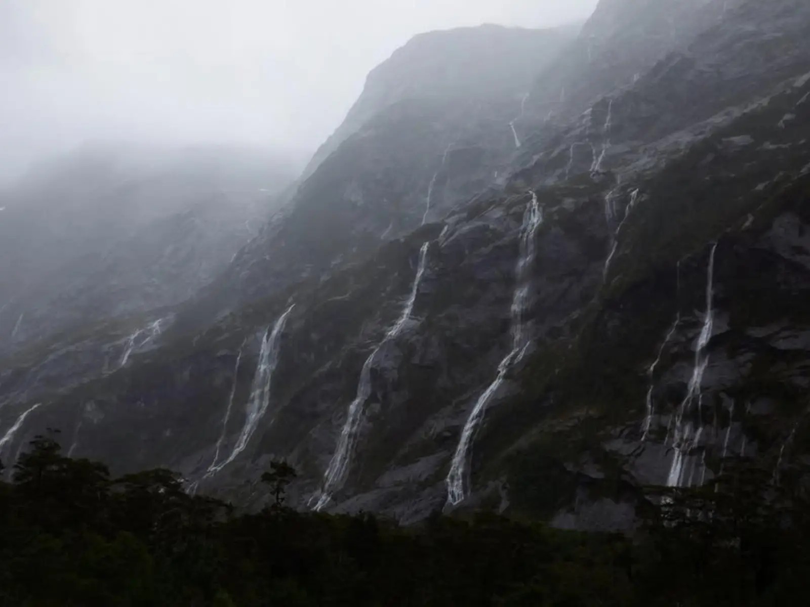 Multiple thin waterfalls cascading down steep, mist-covered rock faces near Homer Tunnel in Fiordland National Park during overcast weather