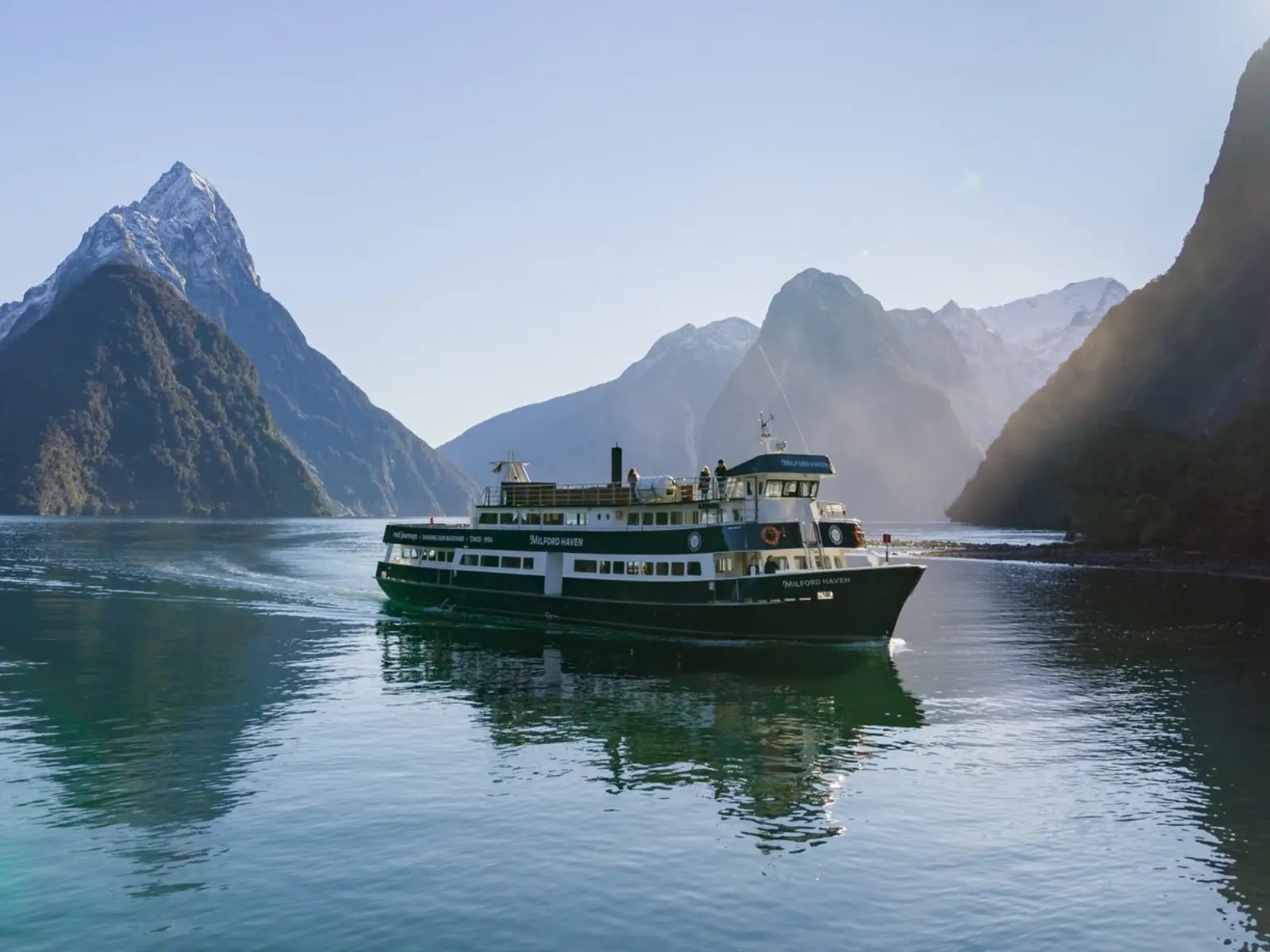 Cruise boat travelling through Milford Sound with calm water and dramatic fiord cliffs rising on either side under clear skies