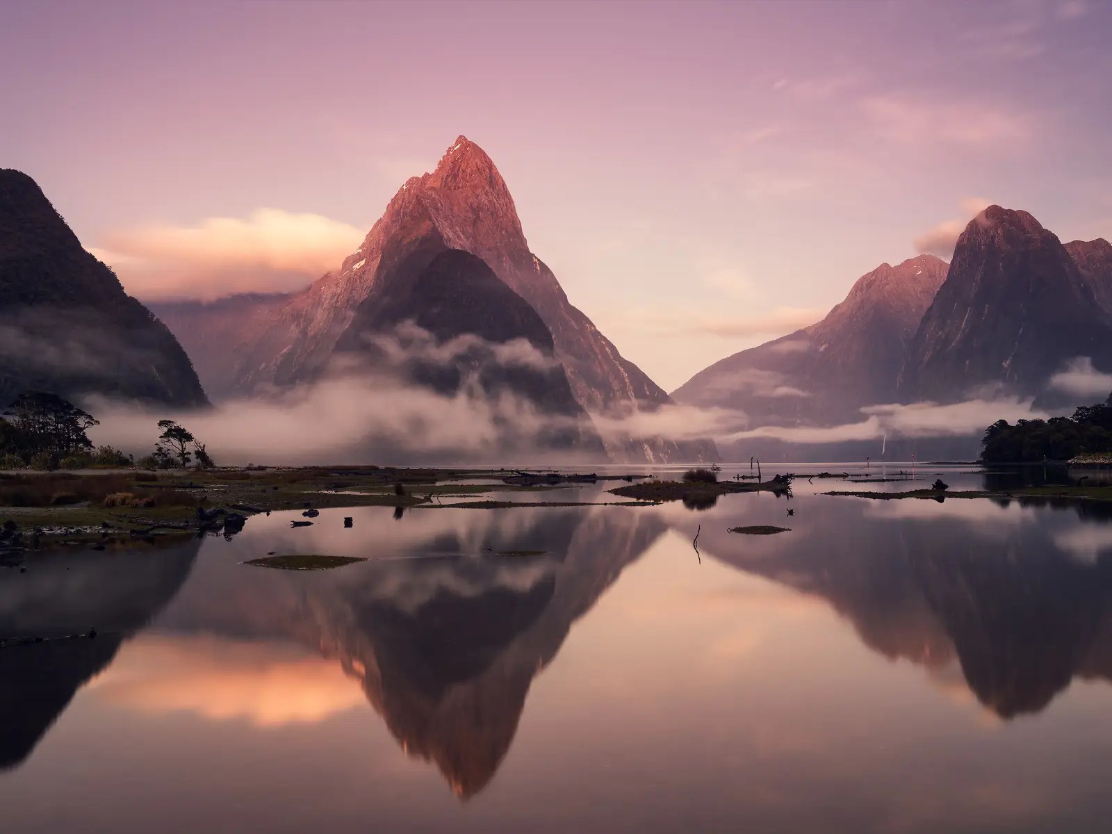 Sunrise over Milford Sound with Mitre Peak reflected in calm water, low cloud drifting through the fiord and warm pink light across the mountains
