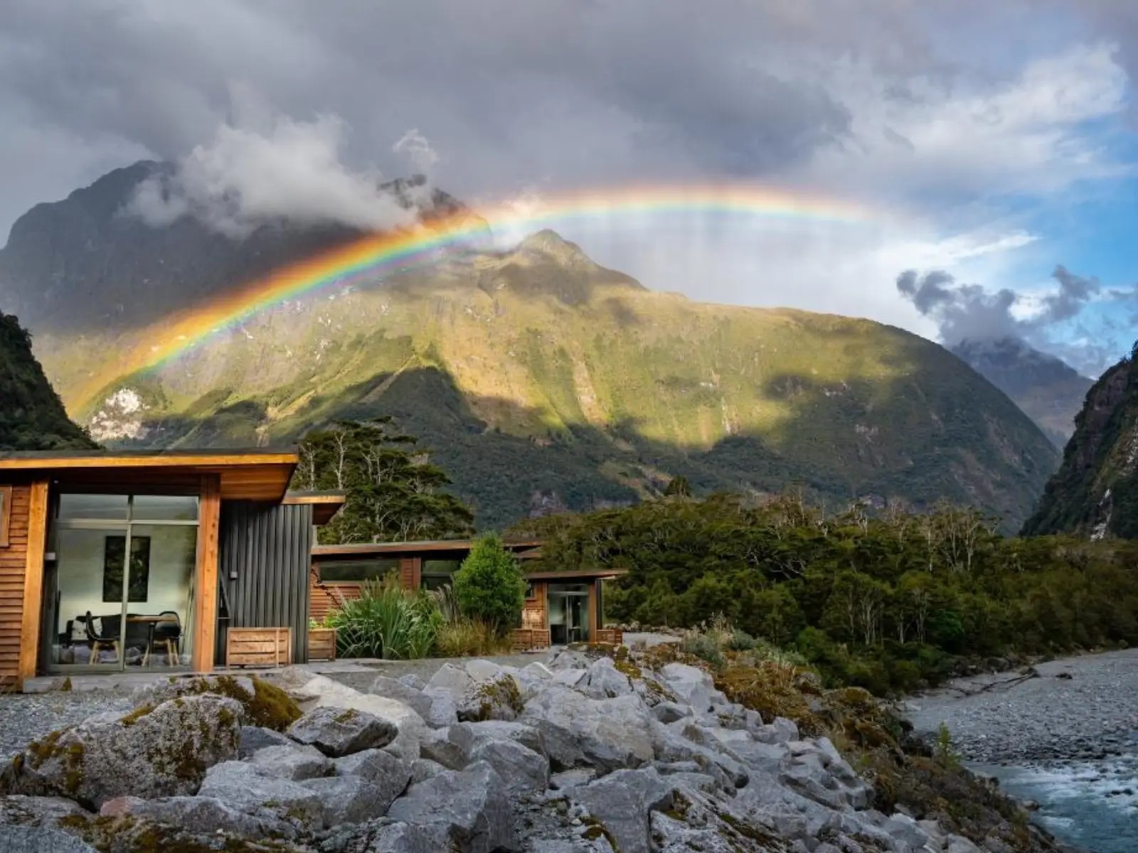 Rainbow over Milford Sound Lodge with modern cabins set beside the river, surrounded by forested mountains and dramatic Fiordland scenery