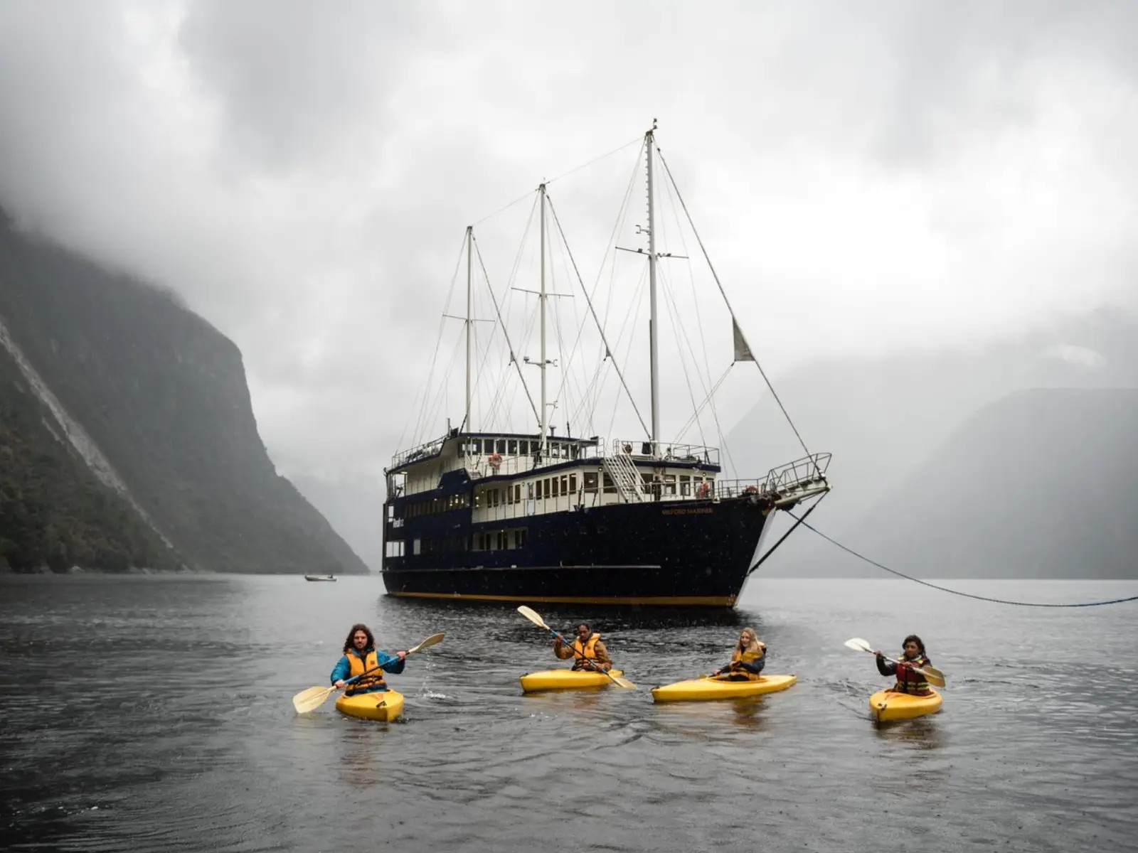 Group kayaking on Milford Sound with an overnight cruise boat anchored behind them, surrounded by steep misty cliffs and calm fiord waters