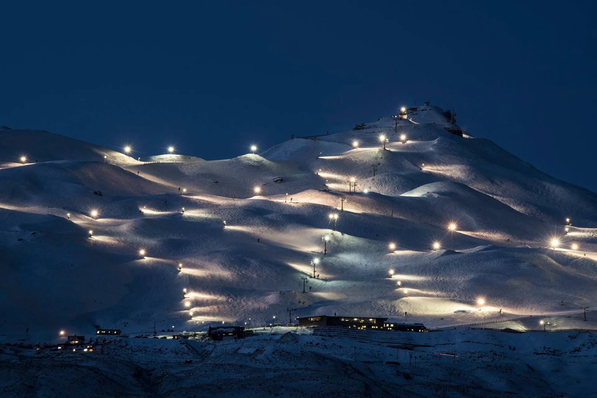 Snowy ski field of Coronet Peak at night, illuminated by dozens of of lights.