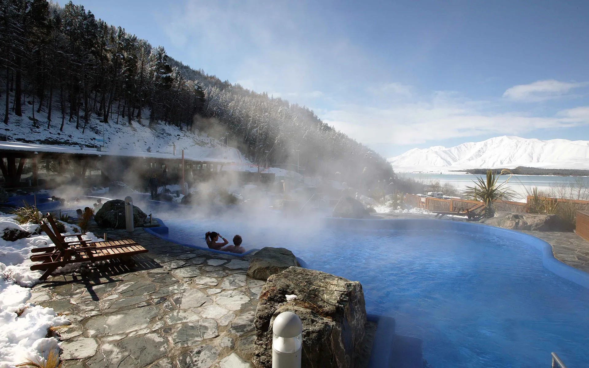 Curved hot pool overlooking Lake Tekapo and snowy mountain forest.