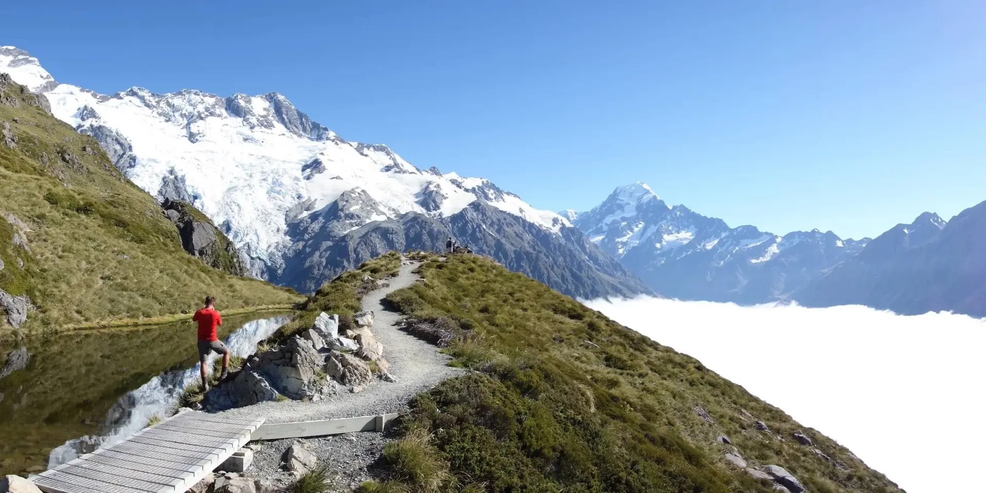 Person hiking a mountain track. River on left of track, clouds on right.