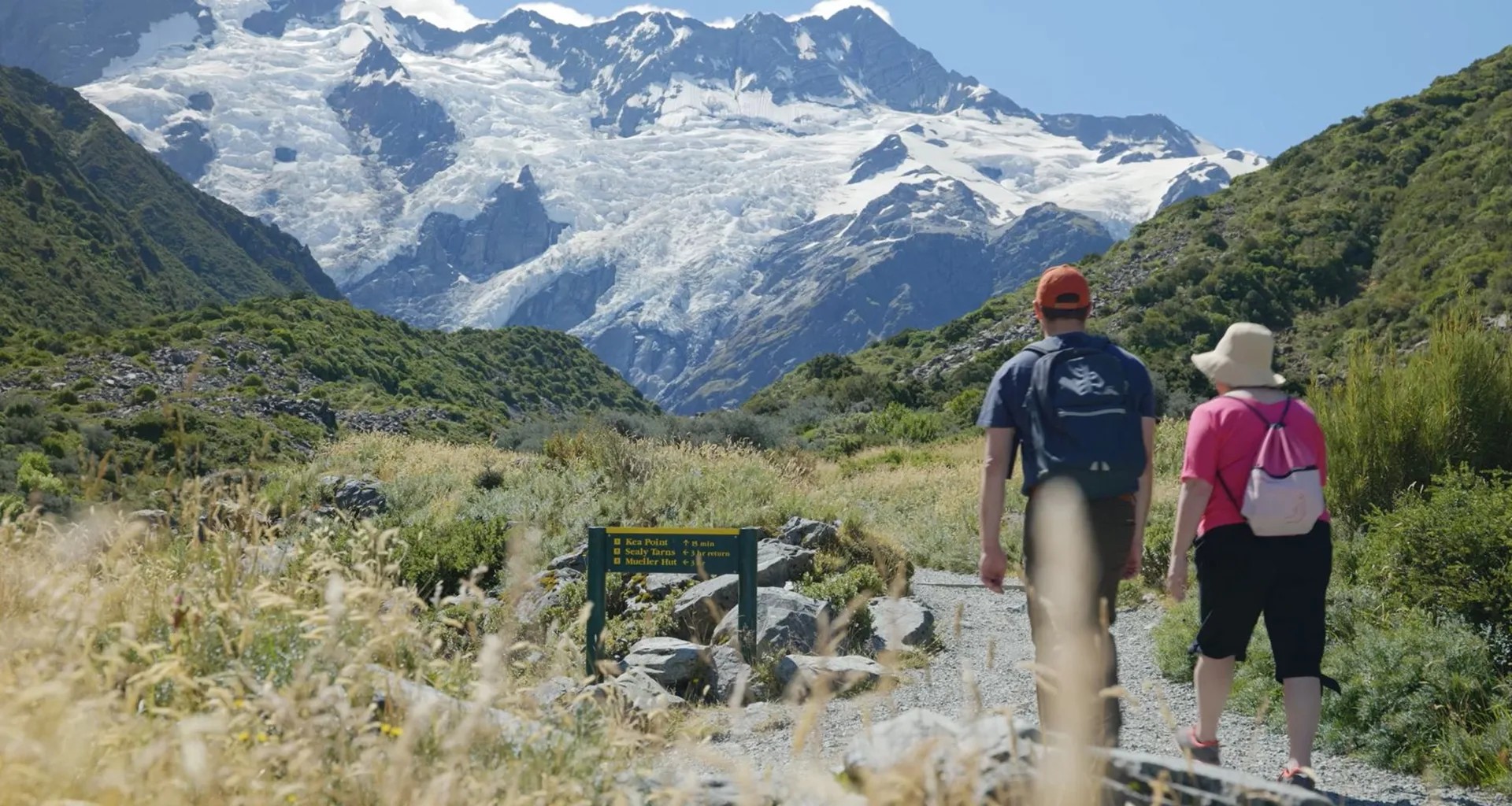 Two hikers walk through a track in a foliage-covered mountain, towards a snowier mountain path.
