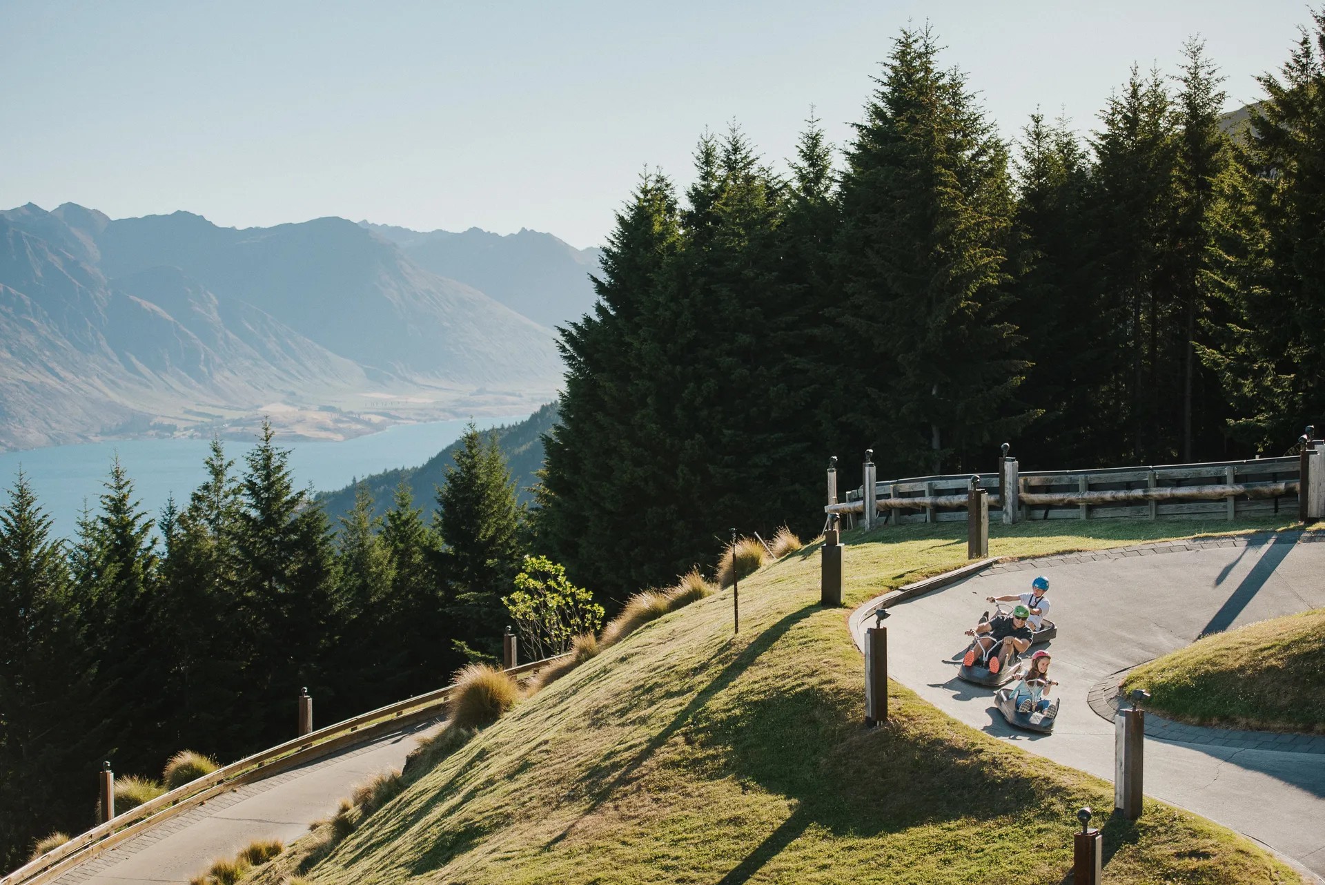 A family rides the Luge, with Lake Wakatipu and surrounding mountains visible in background.