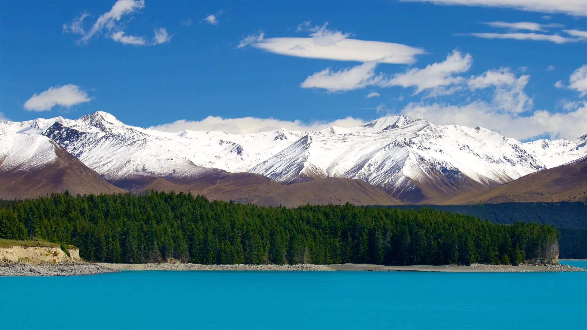 Turquoise lake with an evergreen forest and snowy mountainscape in the distance.