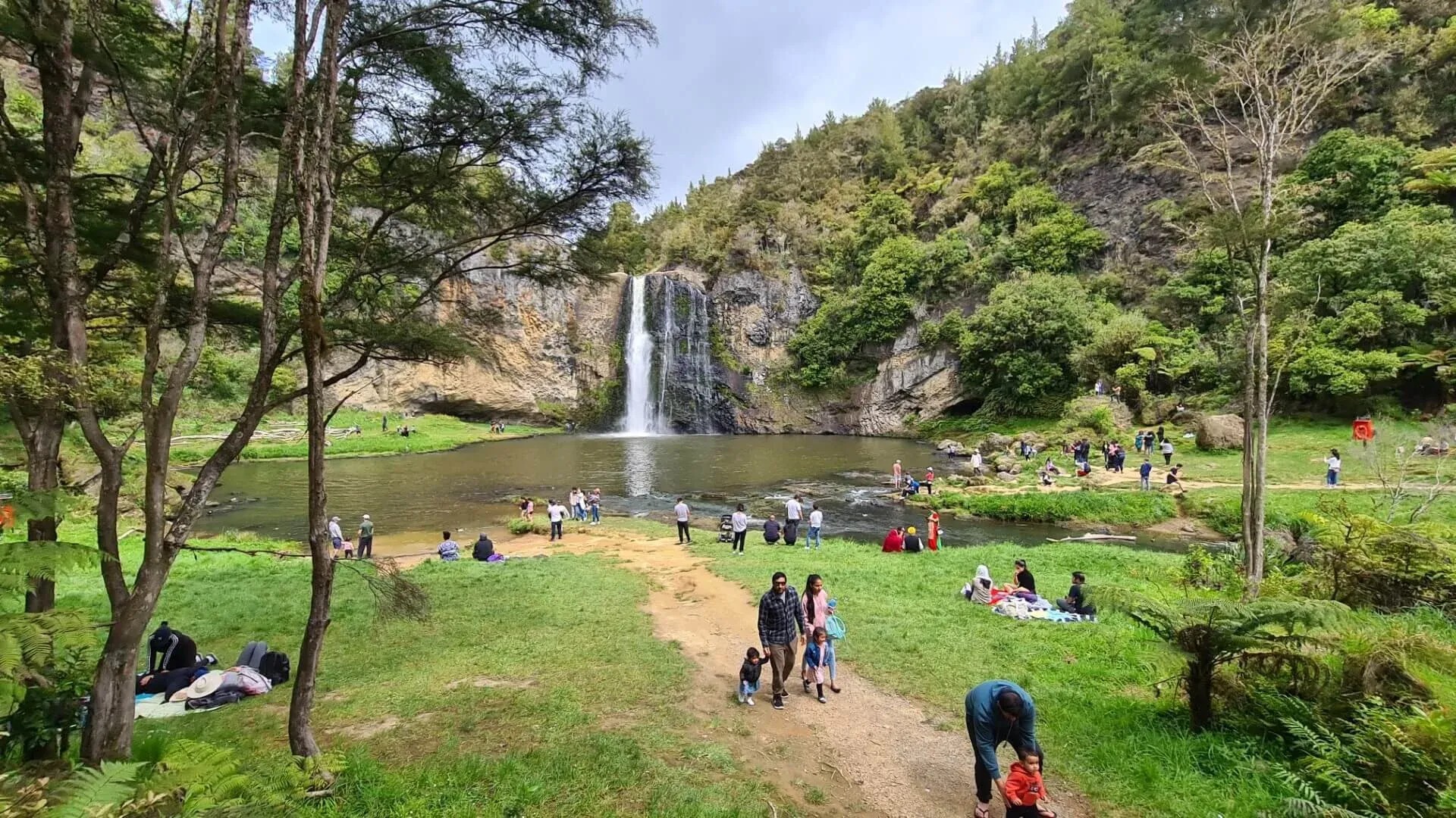 Families walk around a pond and waterfall in a forest range.