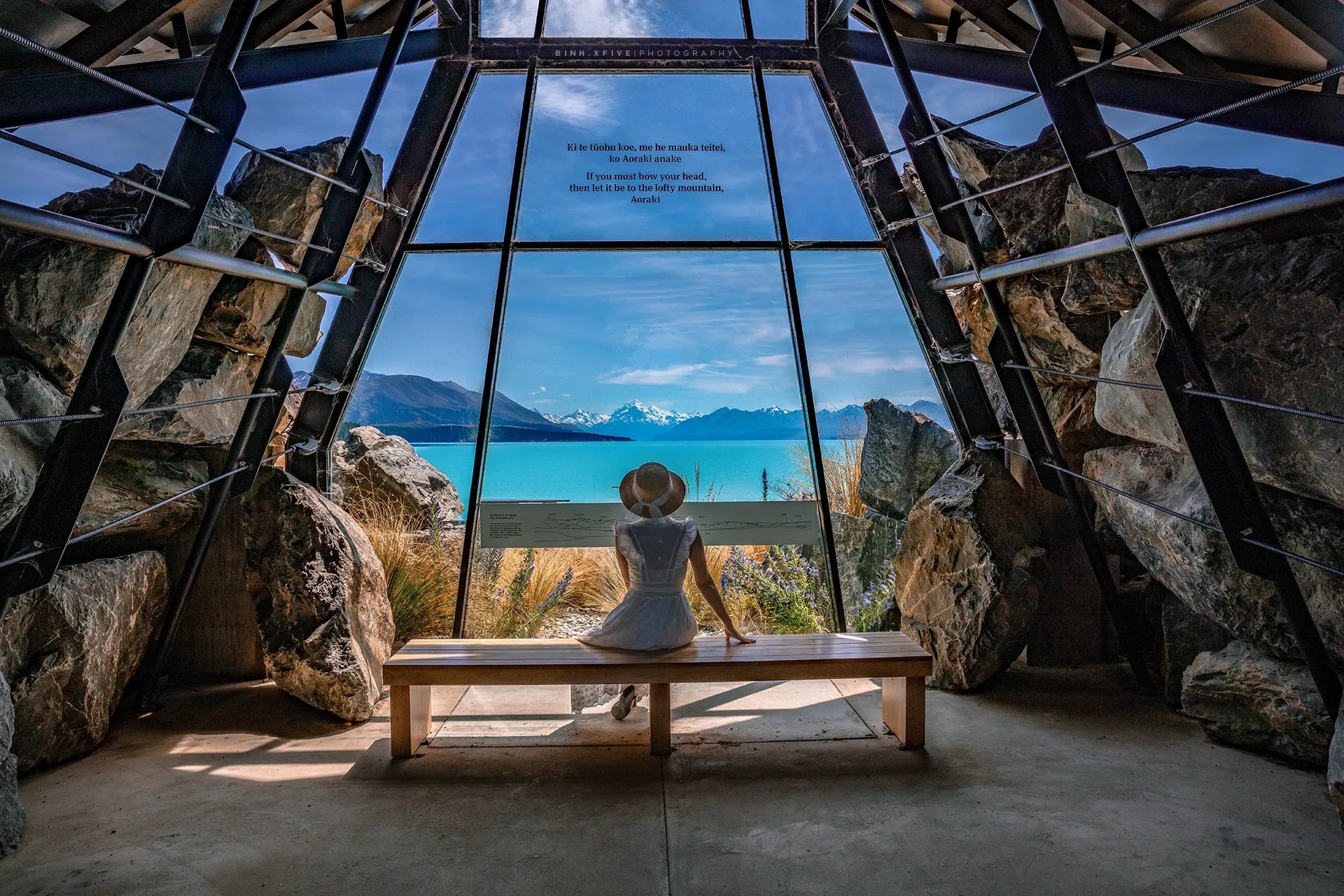 Person sitting on bench in a glass building overlooking a turquoise lake.