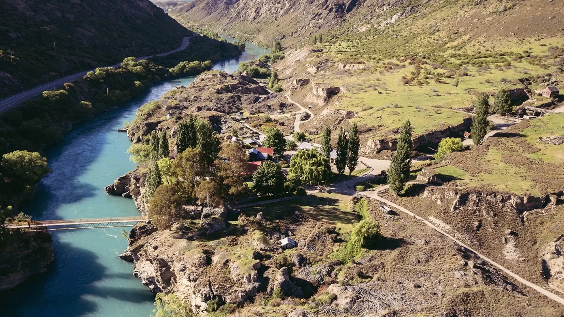 A few buildings sit between a river and a mountain range. Much of the surrounding rock has been mined away.