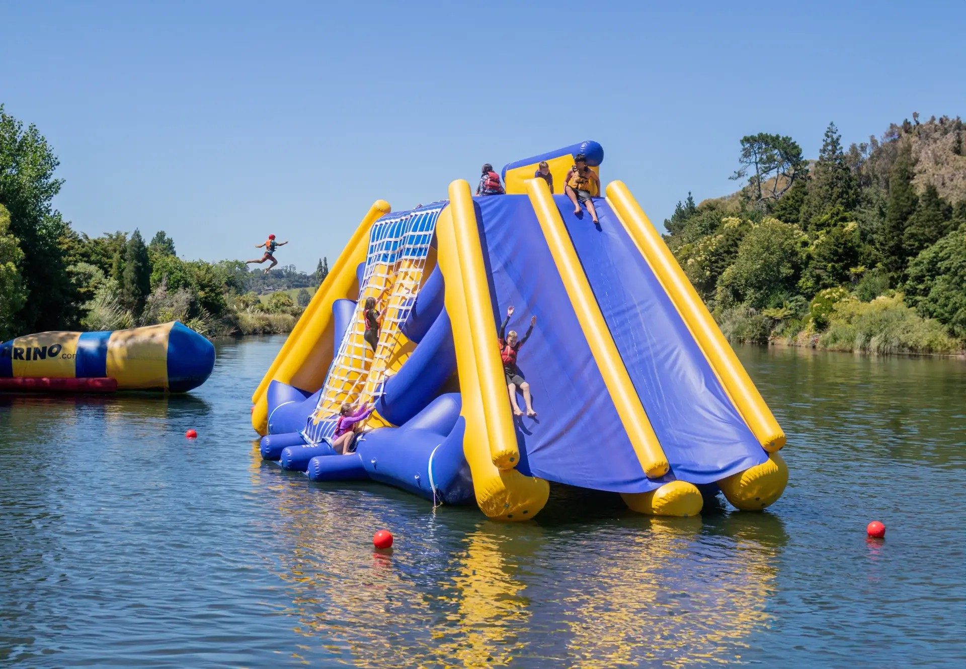 Kids sliding down an inflatable water slide in an outdoor lake at the Waimarino Adventure Park water park.