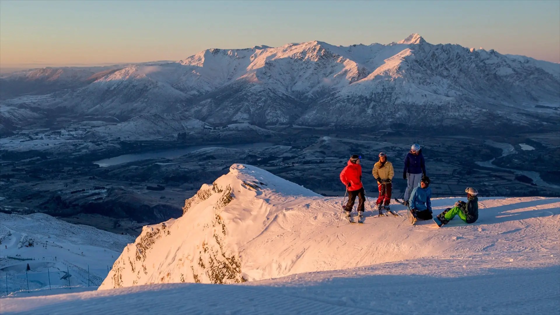 Coronet Peak ski field in winter at sunset, with a group of skiiers and snowboarders resting.