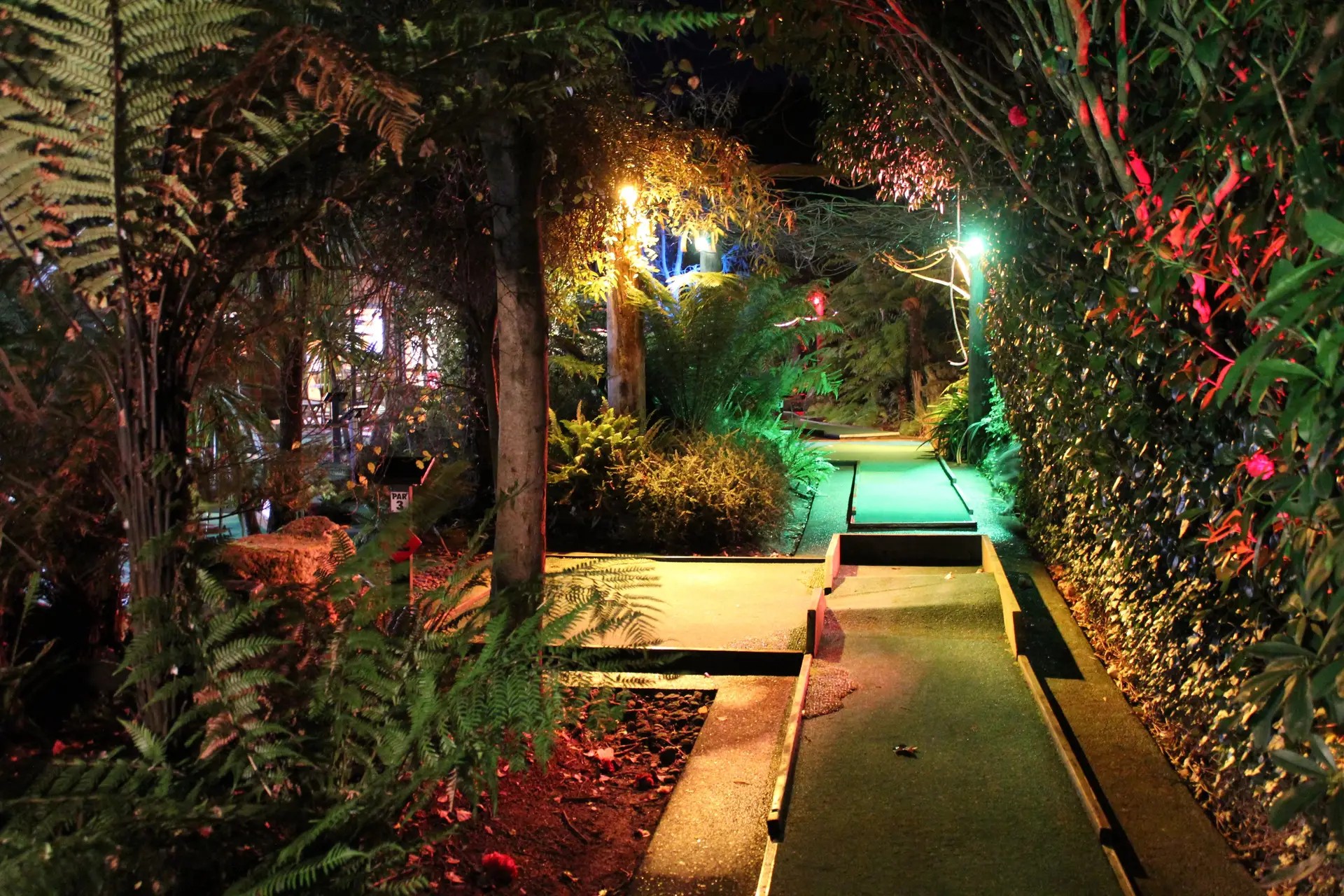 Mini golf course at night, surrounded by foliage and colored lights.