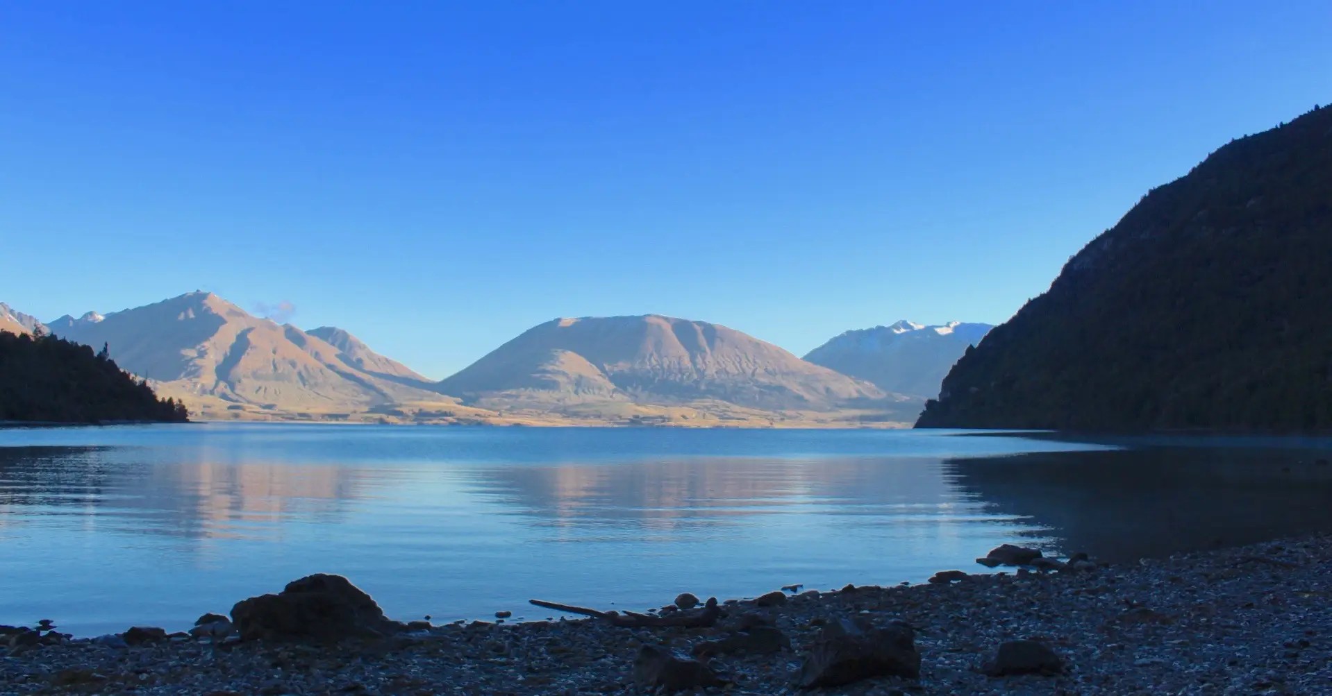 A still lakefront cove surrounded by mountains nearby and in the distance