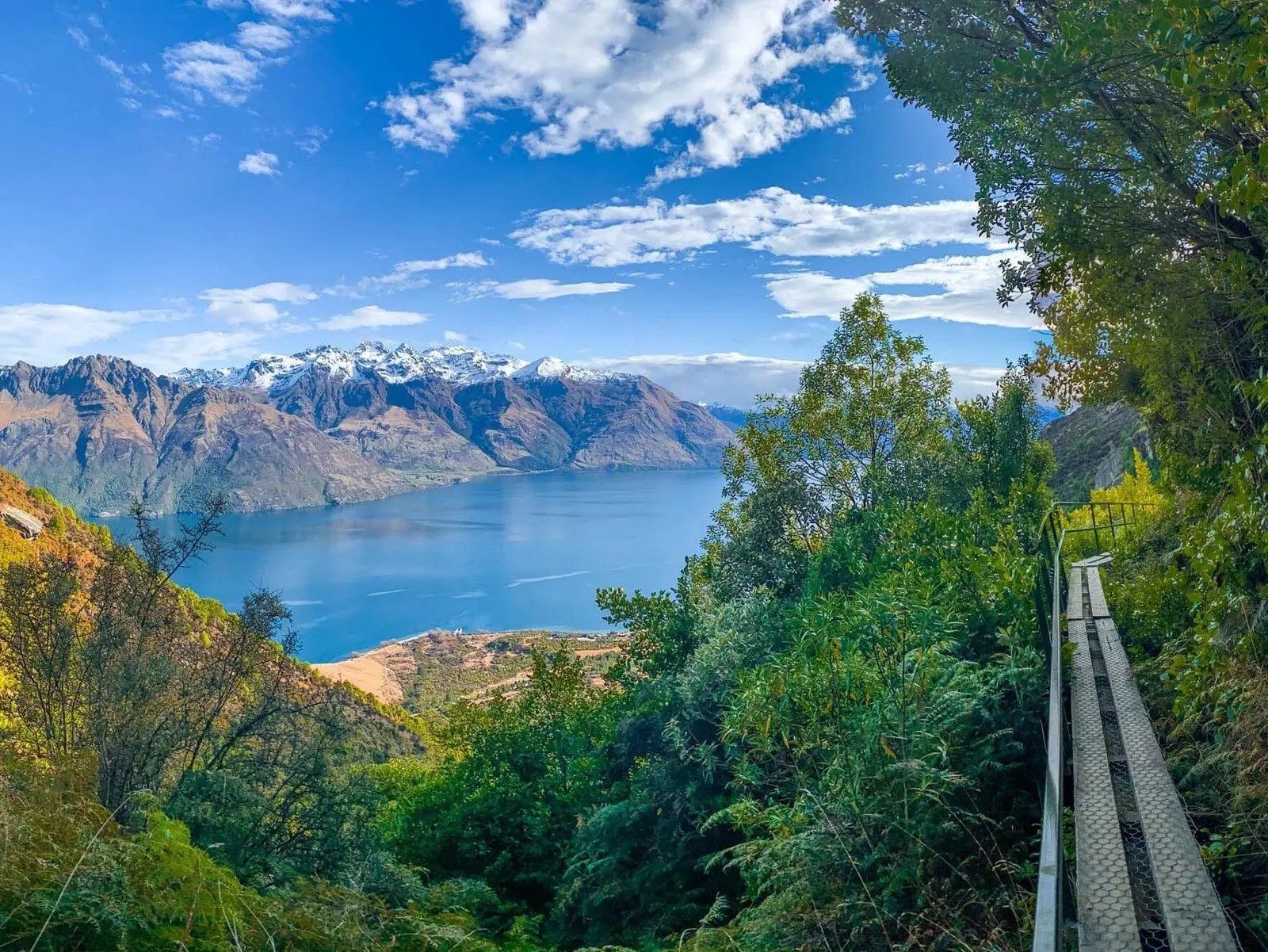 A small path on the side of a hill covered with foliage. The path is made of a metal railing on one side and two sets of wooden planks underneath. The view overlooks a lake and snowy mountains