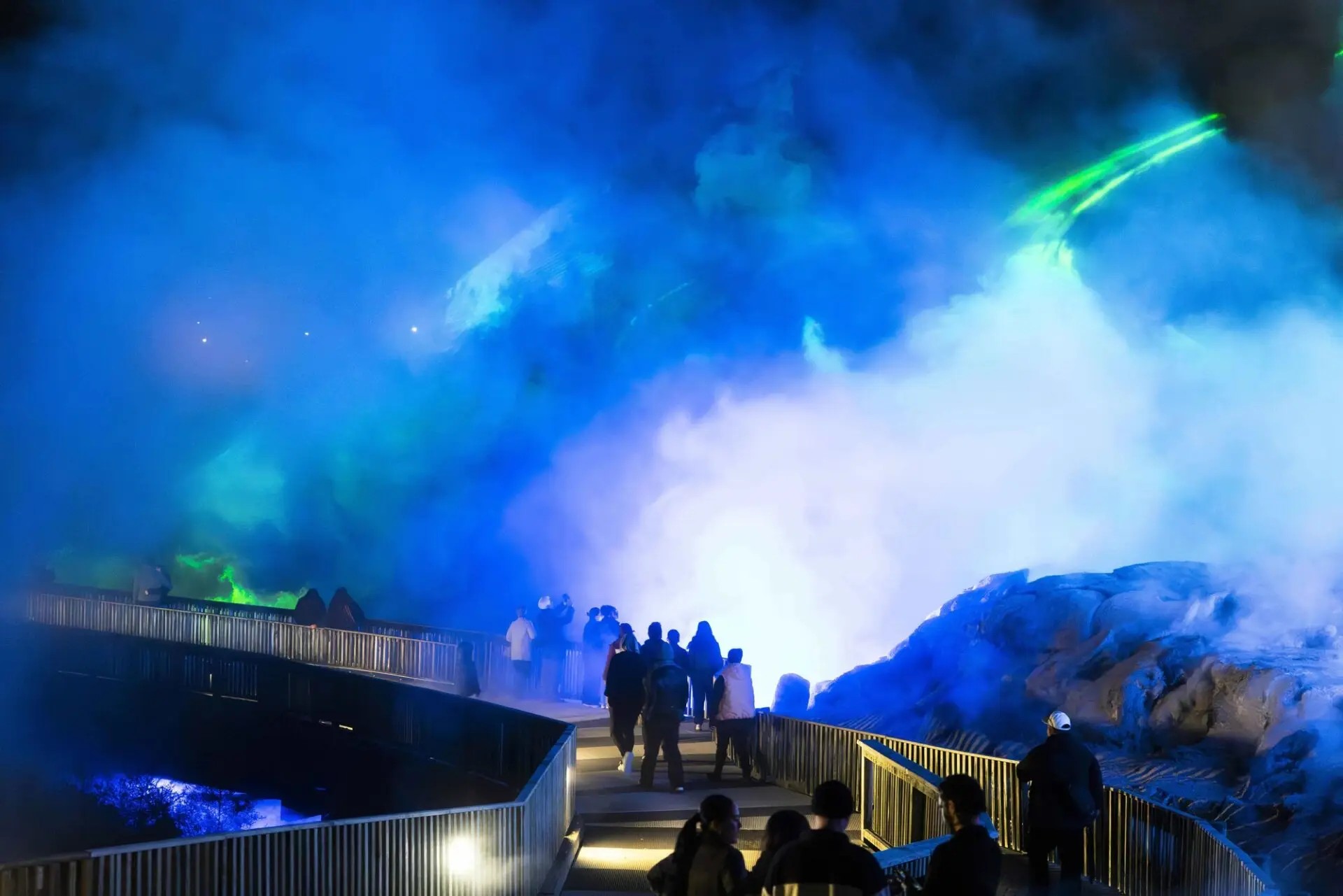 People walking along a wooden boardwalk between geysers lit up by neon green lights