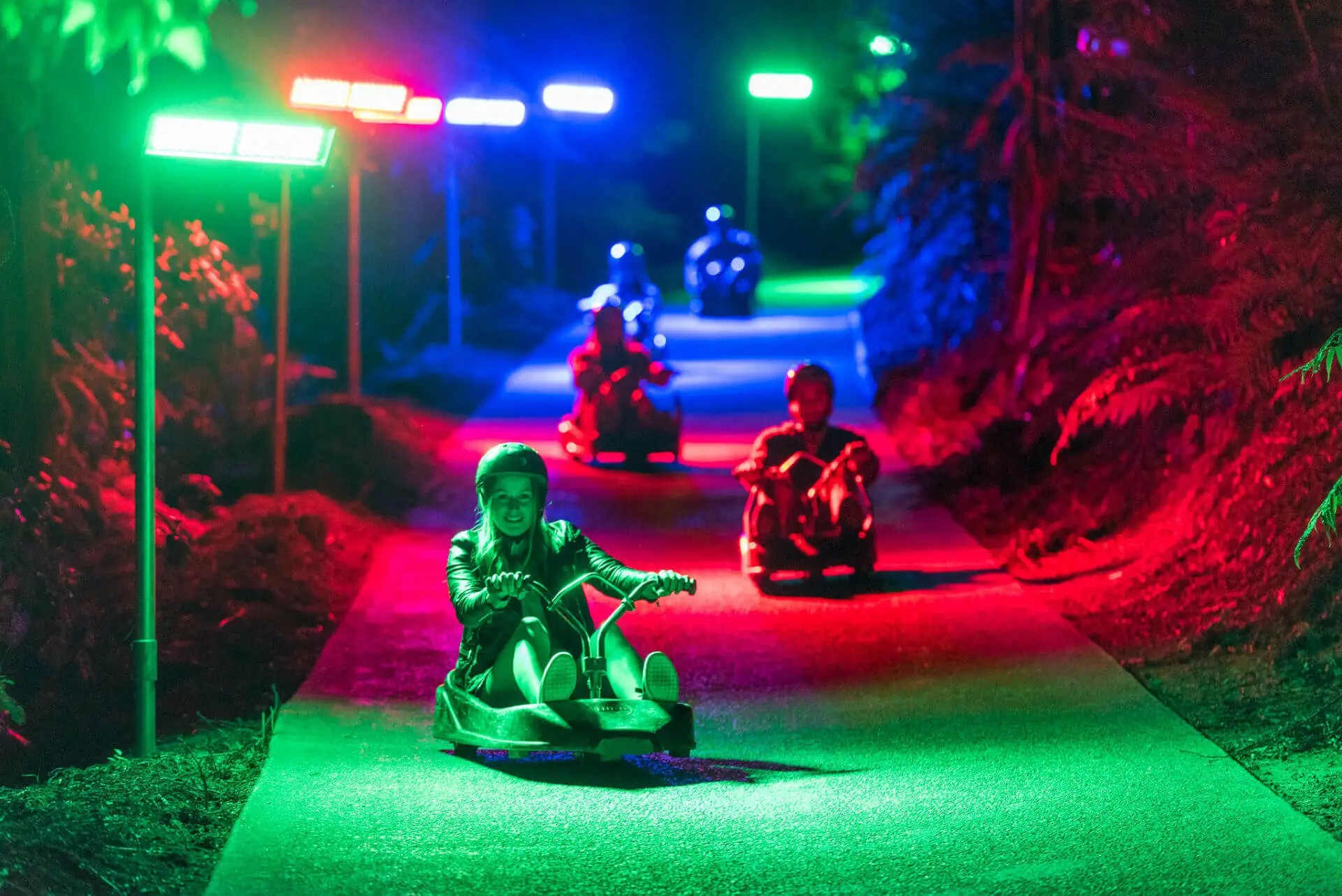 People riding Luge karts down a hill at night, light up by colored lights