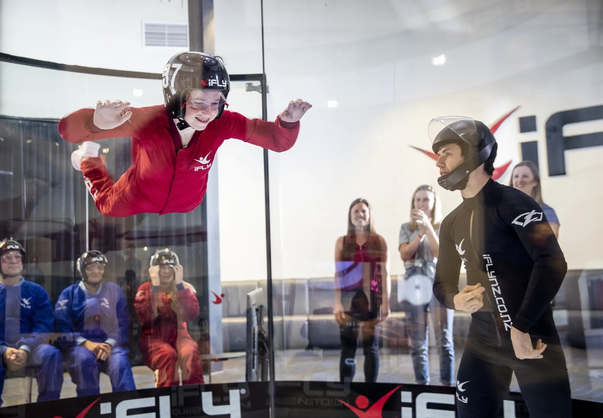 Kid wearing a helmet flying in an indoor skydiving wind tunnel, with an instructor standing nearby and other flyers waiting outside.
