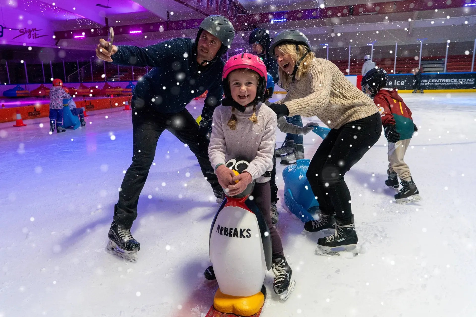 A family on an ice skating rink. The child is holding a penguin-shaped skating aid, while the man takes a selfie and the woman laughs.