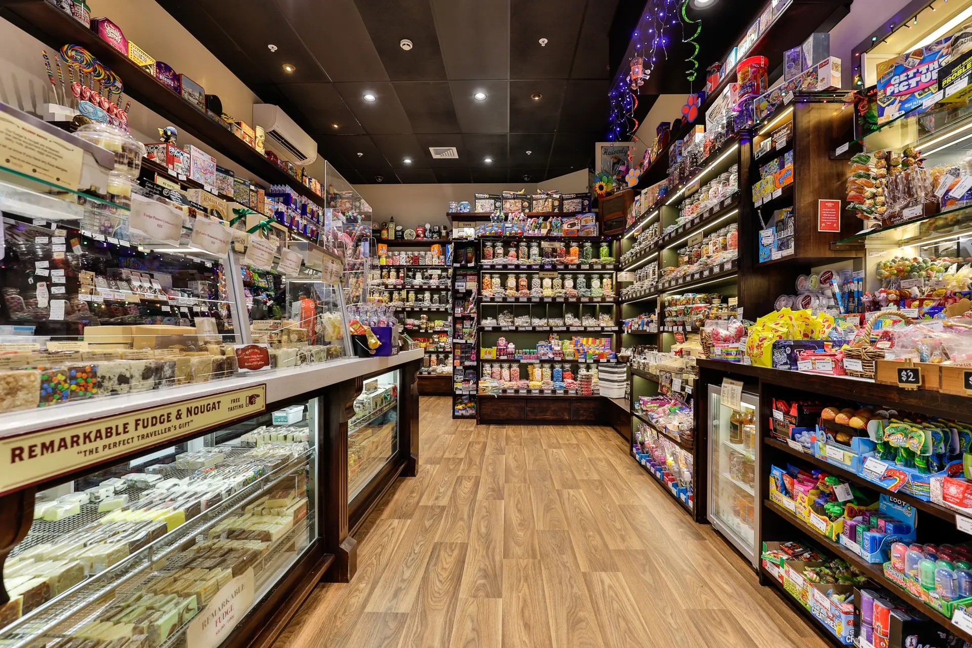 A confectionery shop featuring wooden cupboards filled with sweets. Near the front of the store, the counter displays a fridge full of fudge and nougat.
