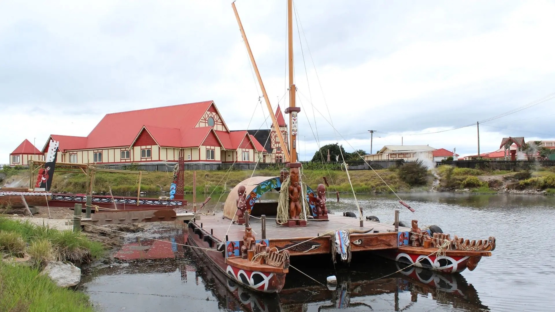 A wooden raft with Maori sculpture and patterns docked in a stream. Behind it is a waka. Behind the stream are some red-roofed buildings and steaming geothermal vents.