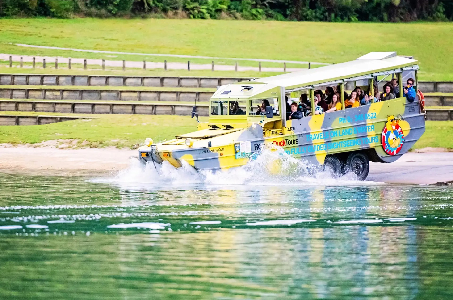 An amphibious military vehicle splashing from land into water. The vehicle has a duck mascot wearing sunglasses.