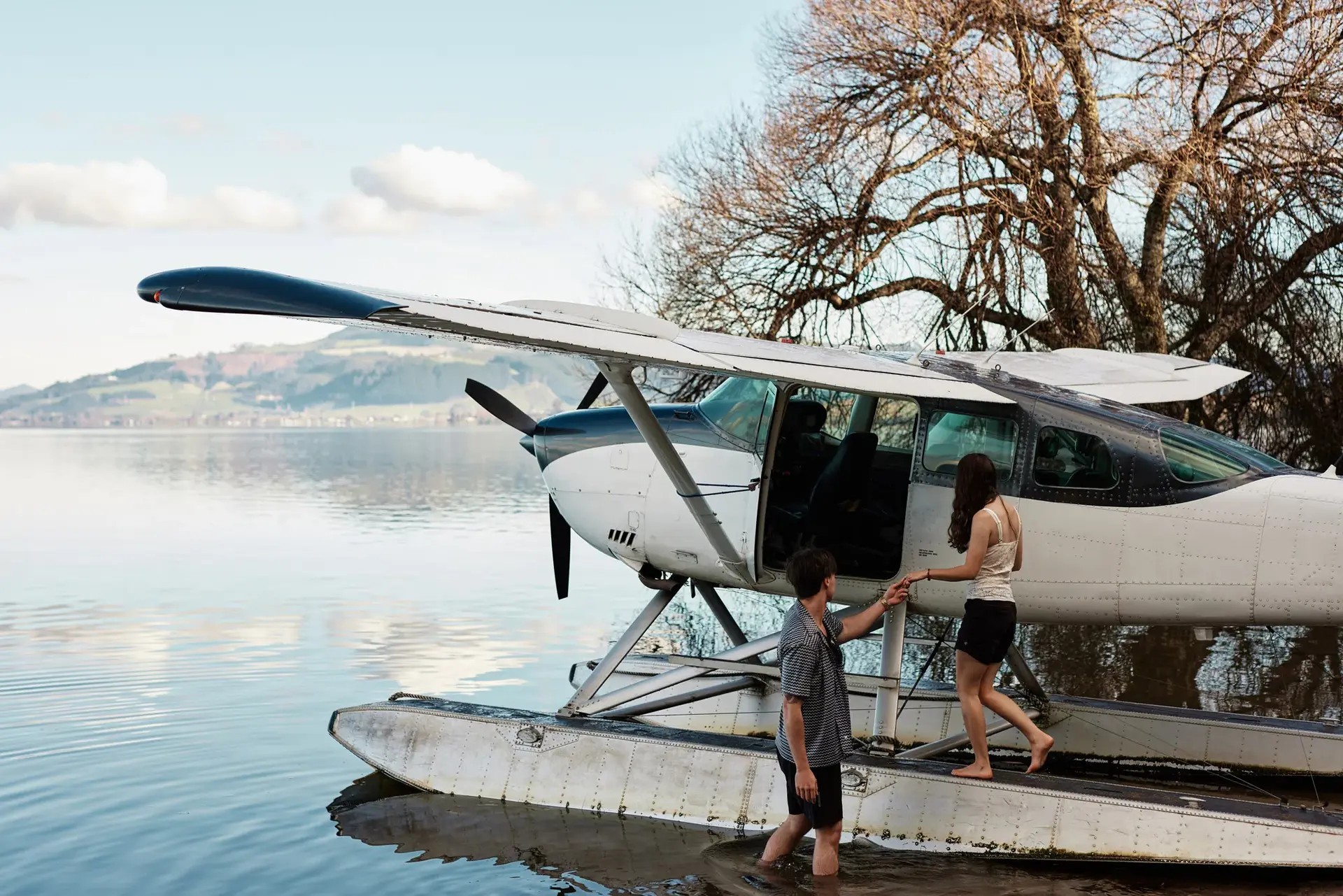 A man and a woman enter a white floatplane by the edge of a large lake. The plane is parked next to a leafless tree growing out of the water.