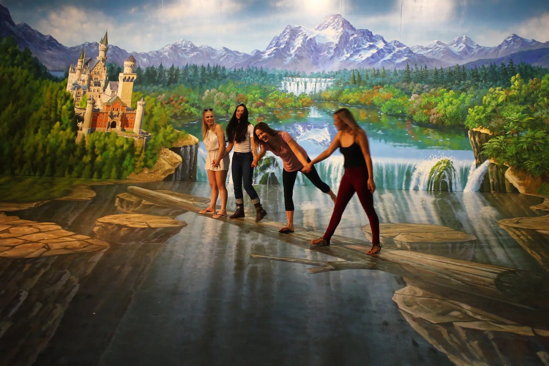 Group of friends holding hands in a trick art gallery room. The perspective of the room's floor and wall paintings makes it look like they are standing on a log above a waterfall.