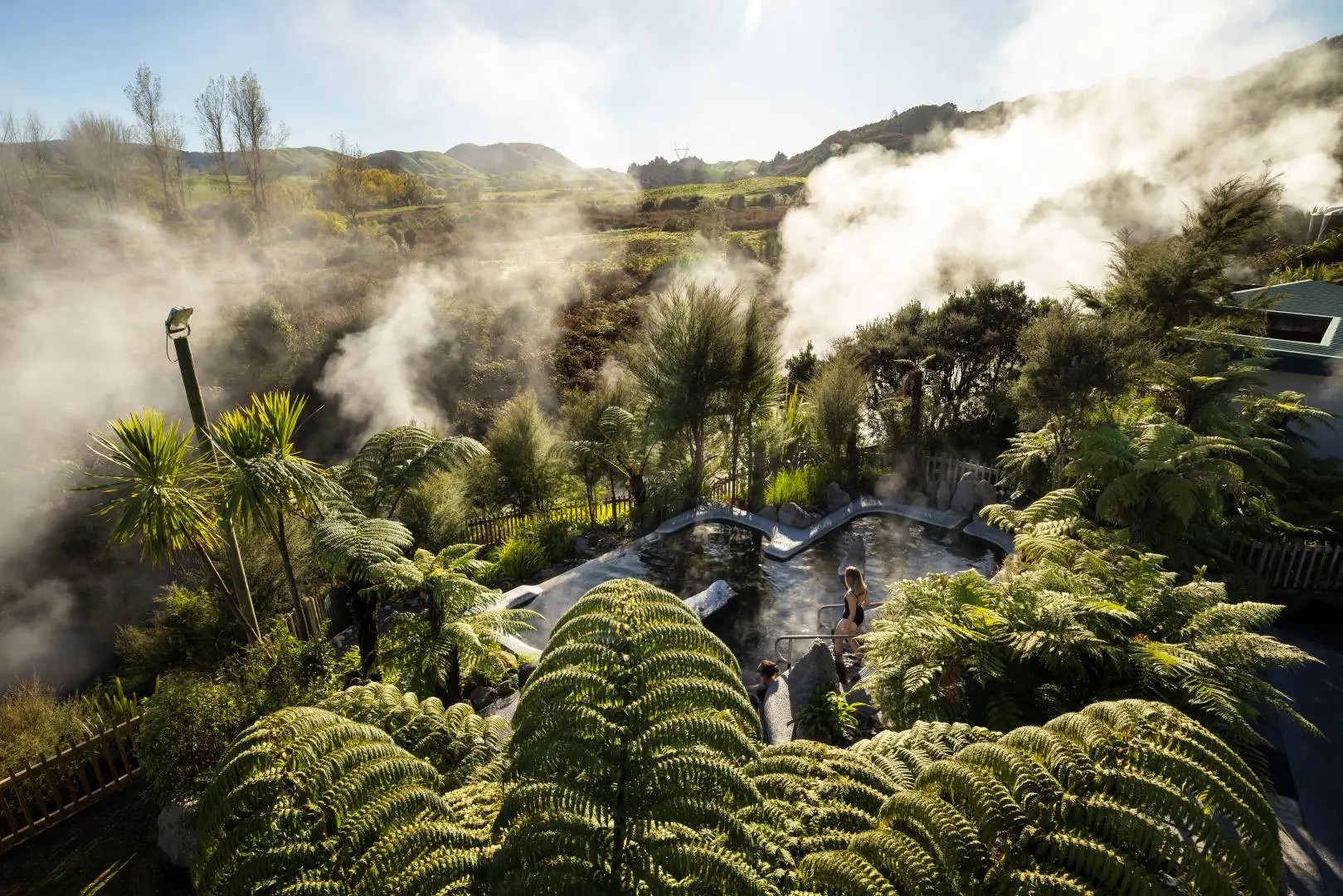 A woman enters a hot pool in a hilly area surrounded by native trees. Steam rises from a nearby geothermal spring.