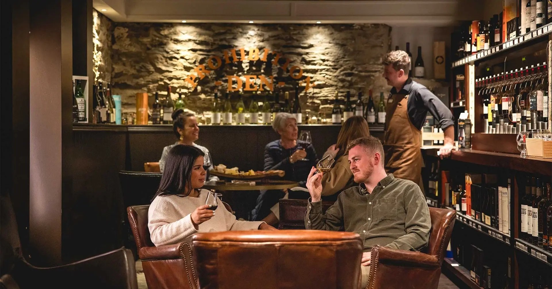 Two people tasting wine while sitting in a small bar. There are many bottles of wine on the shelves. Behind them, there is a table with a cheese platter, where more people are tasting wine.