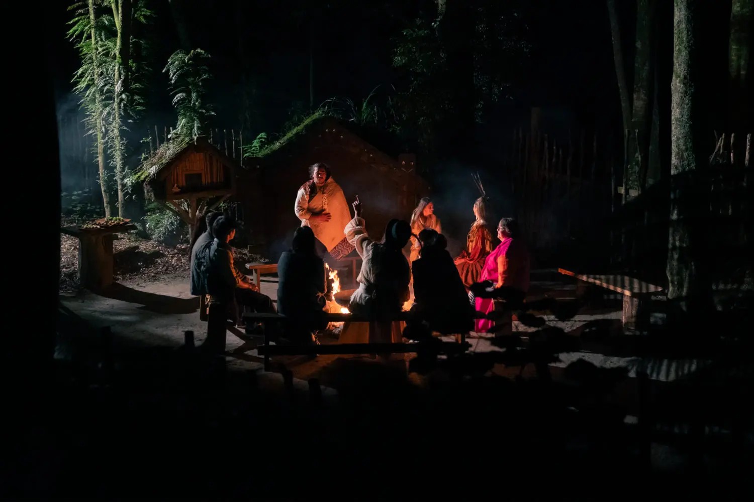 A group of people inside a forest clearing, lit only by the fire in the centre. Some of the people, dressed in a mix of traditional Māori clothing and European dress, are telling a story while the others listen.