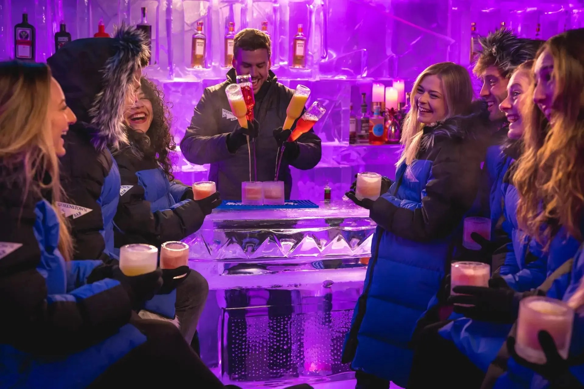 A bar where the counter and shelves are made of ice. The bartender is making a cocktail, while customers, dressed in parkas, are laughing and enjoying their drinks.