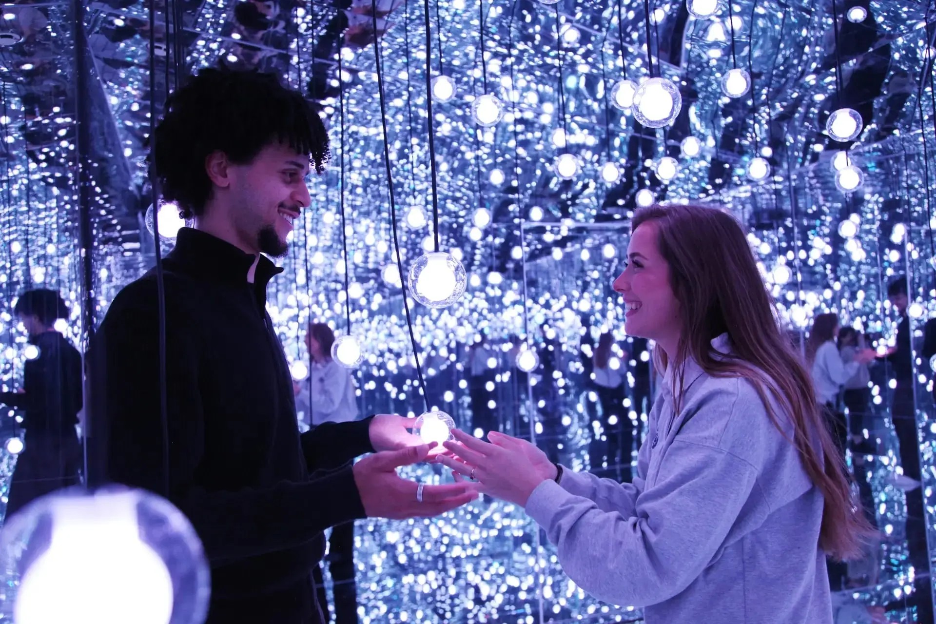 A room where each side is covered in mirrors. There are many round lightbulbs hanging from the ceiling. A man and woman smile at each other while holding one of the lightbulbs.