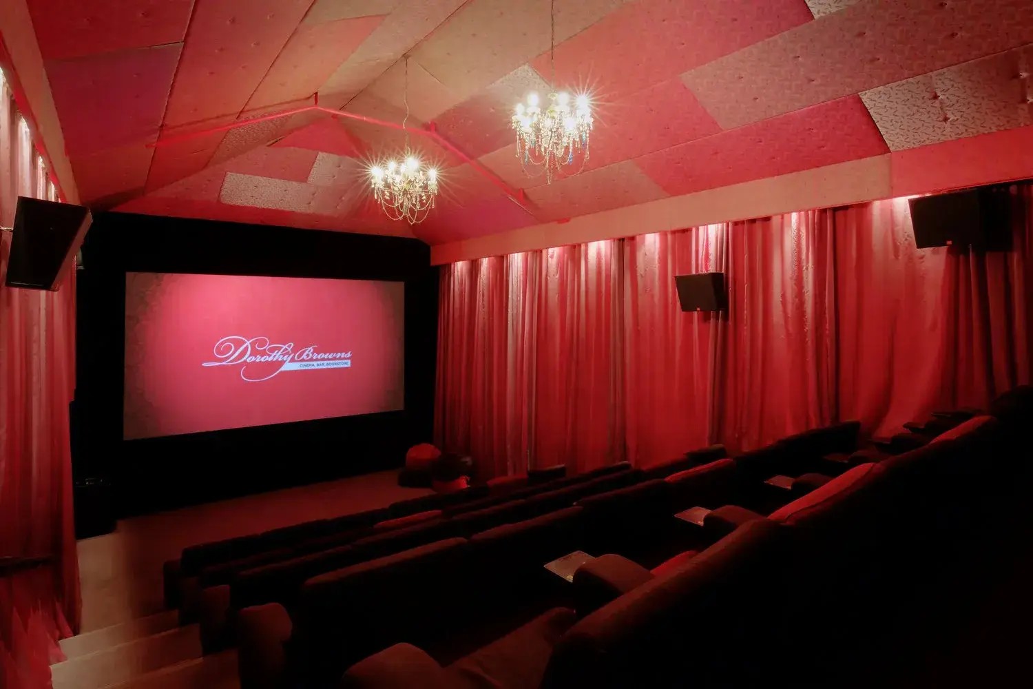 A small vintage-themed movie theatre with red plush chairs, red curtains,a nd pink ceiling insulation.