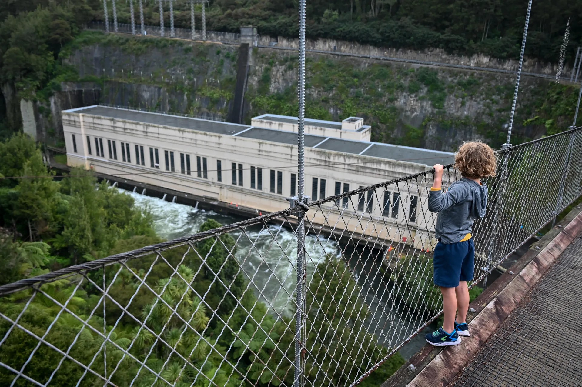 A child stands by the edge of a suspension bridge, holding onto the rail. He looks over the river and hydro power station below.