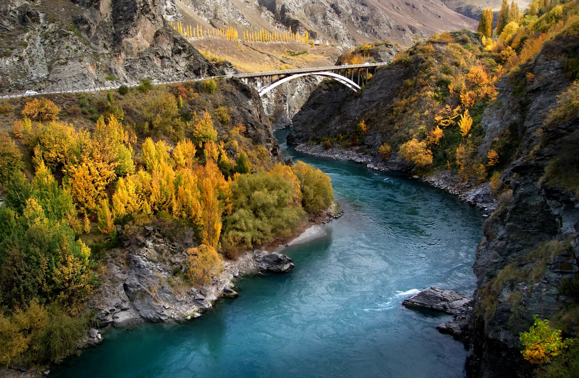 A large turquoise river between rocky, mountainous formations. The trees are yellow-green and there is a large bridge over the river.