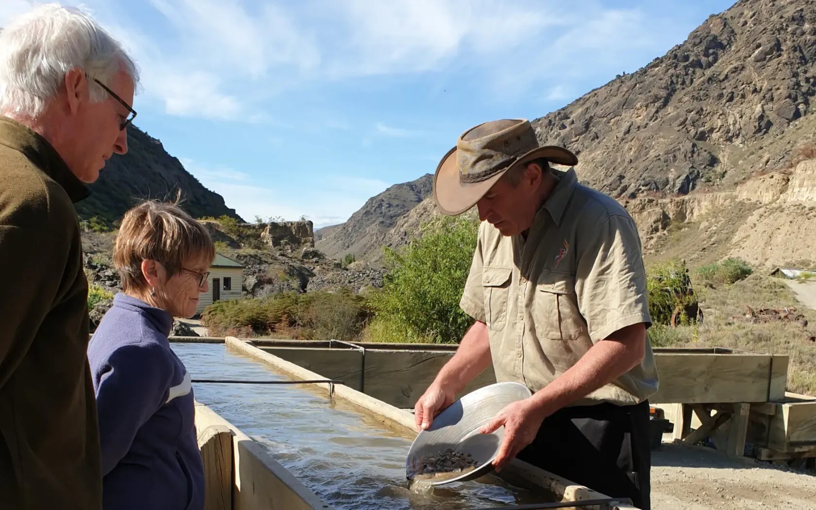 Man shows two people how to pan for gold in a wooden trough filled with murky water.