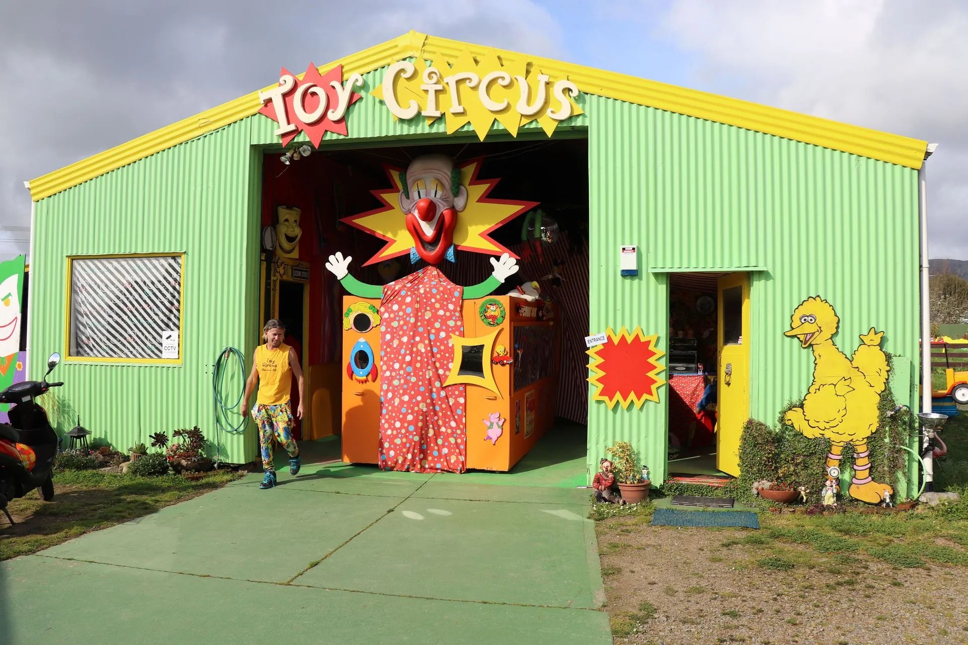 Green corrugated iron warehouse building with a fun sign that reads "Toy Circus". Inside is a structure with a clown head. Outside is a cutout of Big Bird.