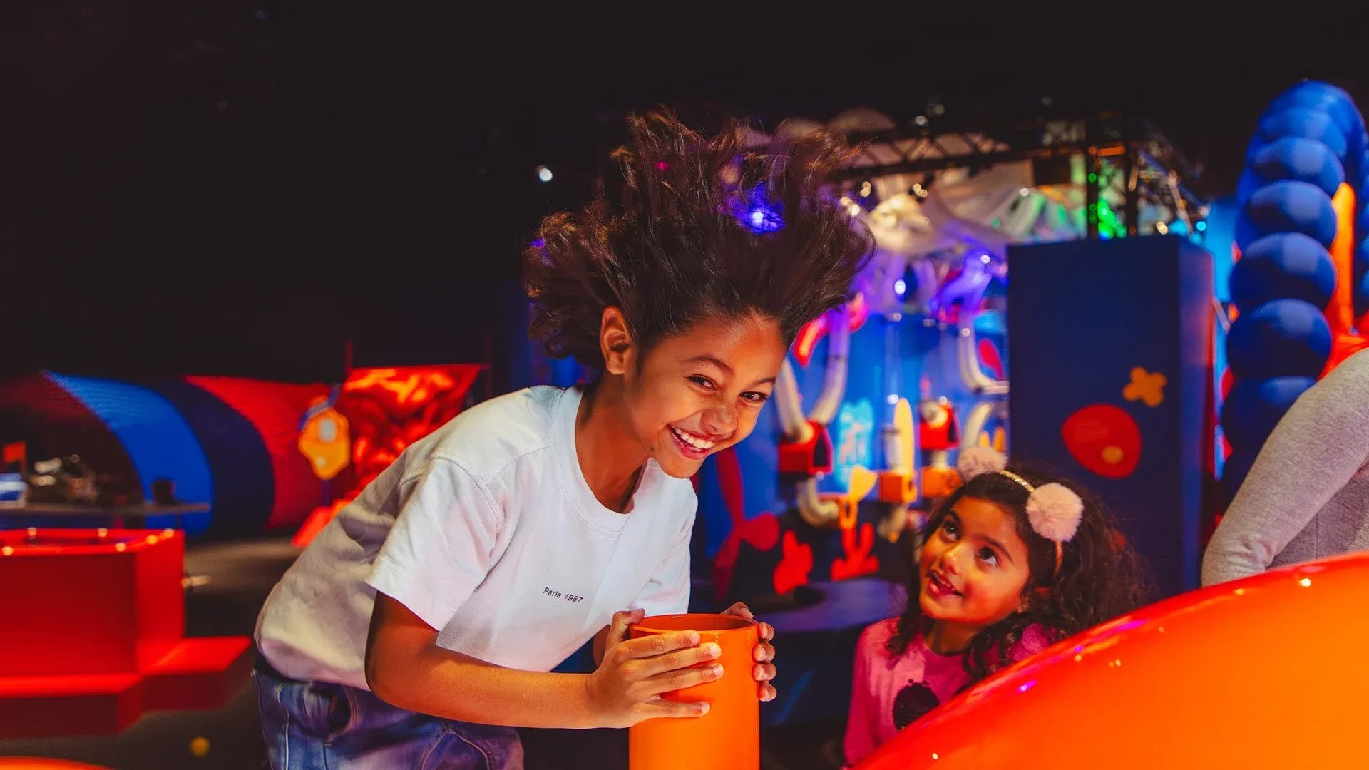 Museum exhibition with big colourful devices. A kid is in front of a pipe which is blowing air into her face, lifting her hair.