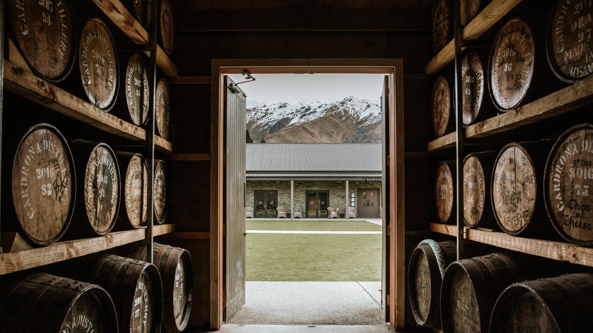Interior of a cellar with wooden casket barrels. Door is open, looking at stone buildings across a grassy courtyard. Behind are snow-peaked mountains.