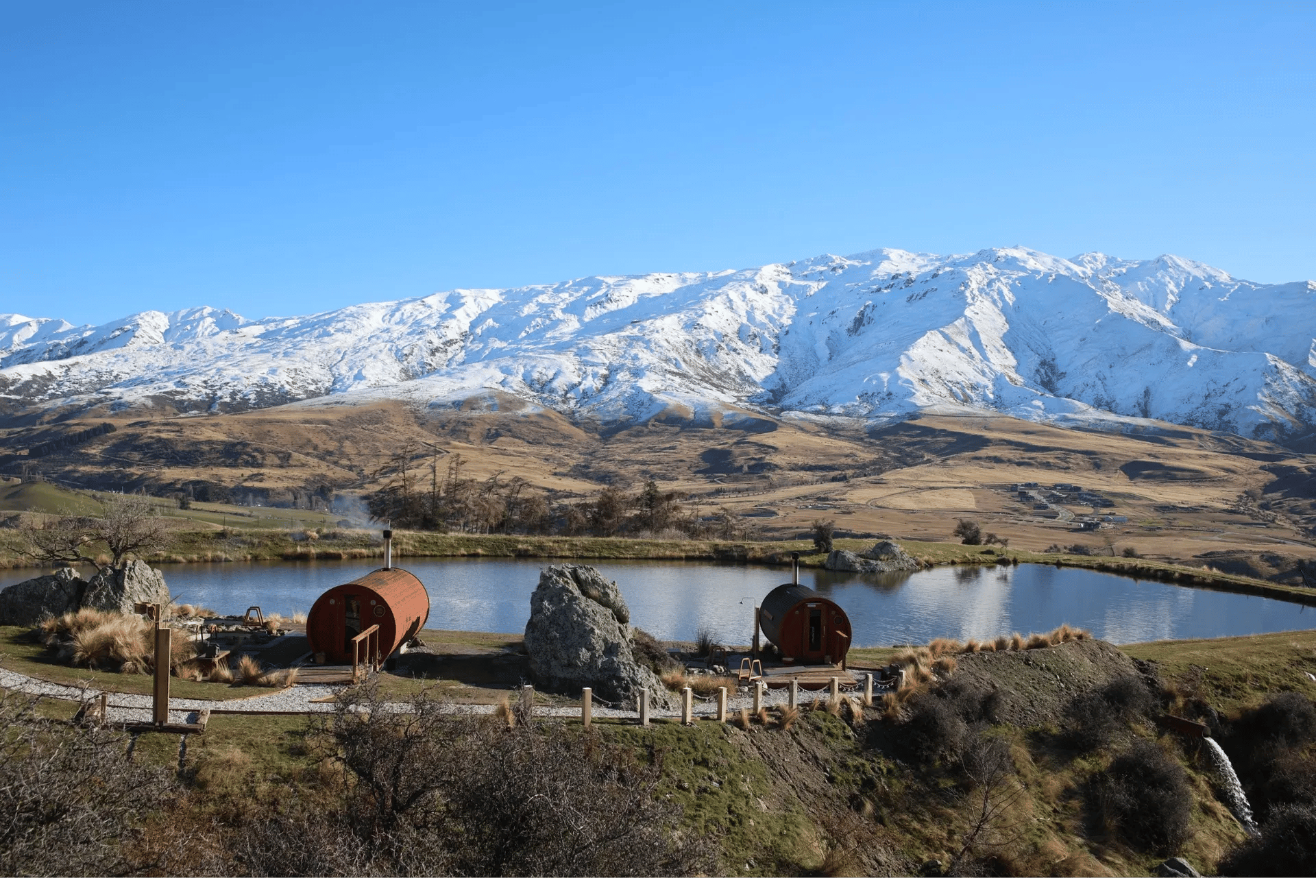 Two wooden sauna barrel-buildings overlooking a small lake and snowy mountain range.