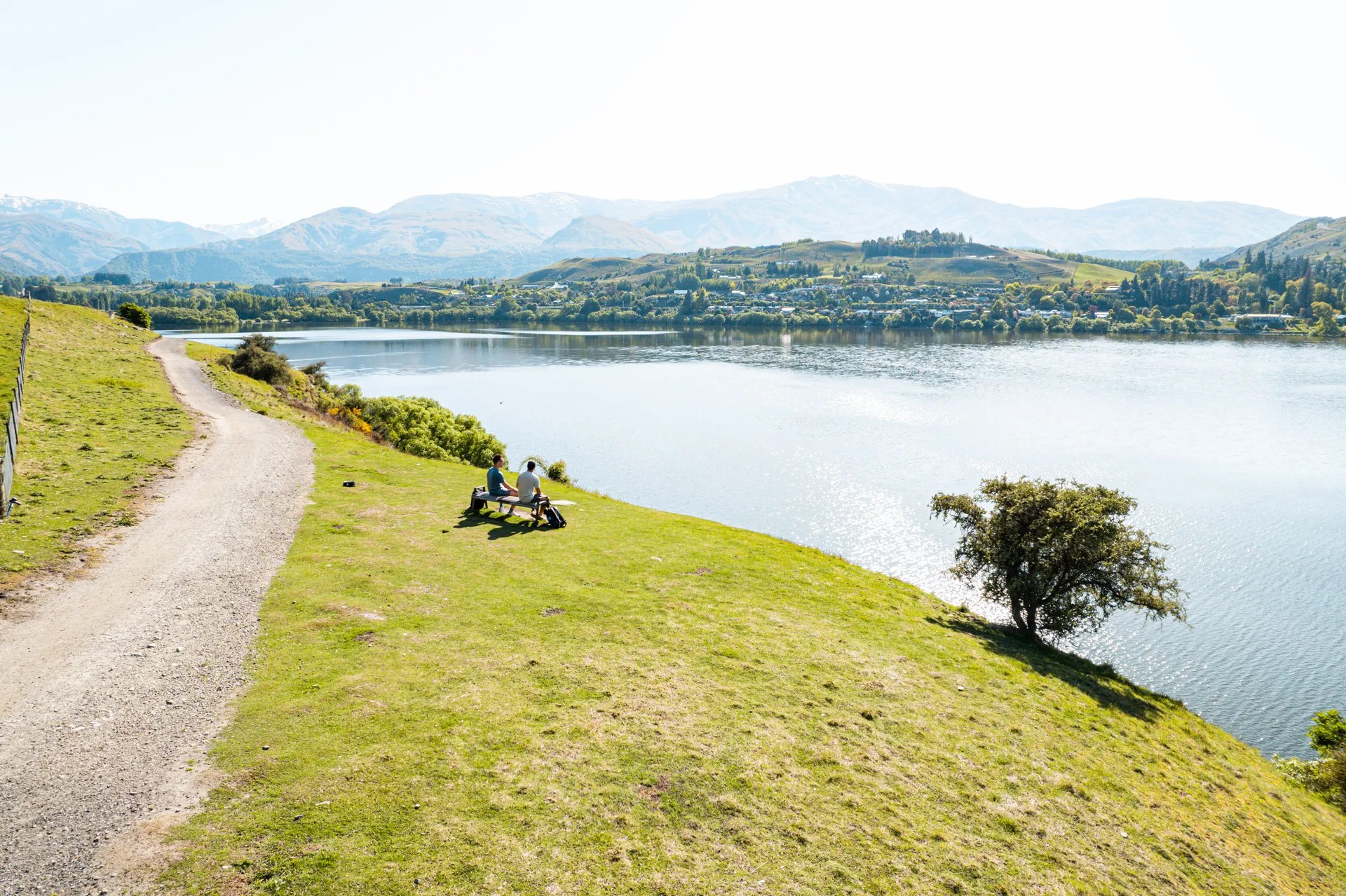 Two people sit on a bench beside the walkway around Lake Hayes.